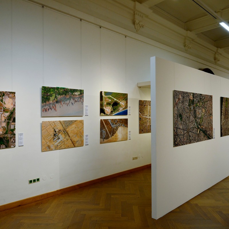 Exhibition displaying aerial photographs of landscapes and urban scenes mounted on white partition walls in a museum
                                 room. (AI-generated alt-text, generated with GPT-4.1-mini)