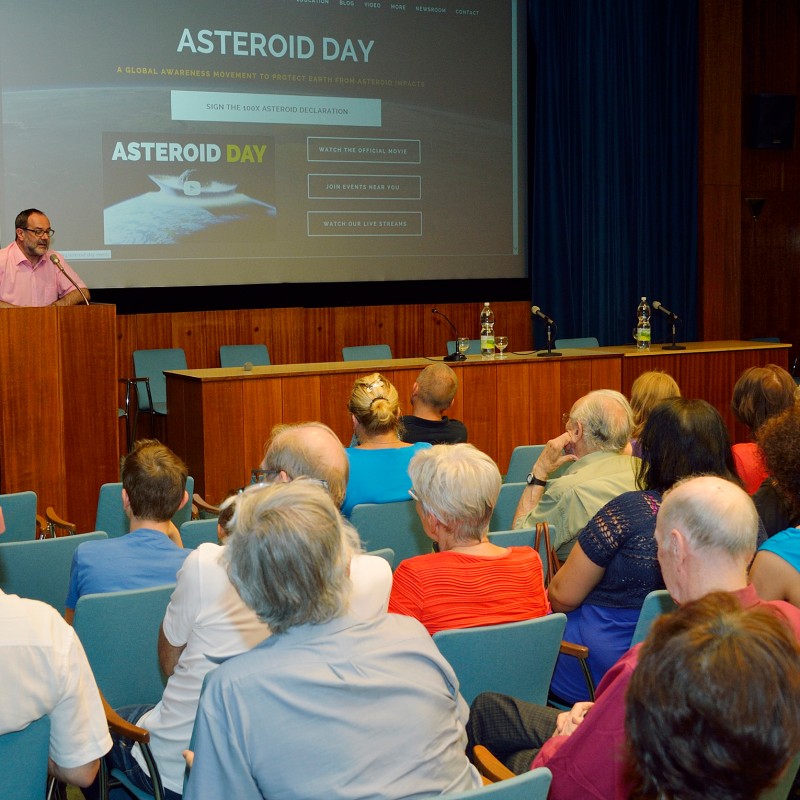 Man in pink shirt speaks at podium to seated audience, presentation with the title 'Asteroid Day' on screen. (AI-generated
                                 alt-text, generated with GPT-4.1-mini)