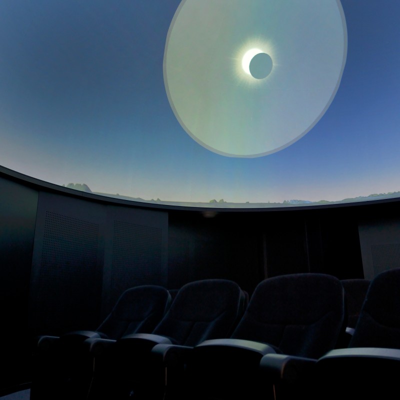 Row of dark-upholstered seats beneath a dome with a projection showing a partial solar eclipse. (AI-generated alt-text,
                                 generated with GPT-4.1-mini)