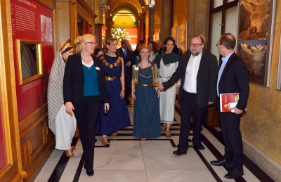 : Mehrere Frauen in historischen Gewändern und mehrere Männer in Anzügen gehen gemeinsam durch einen Museumsflur mit Ausstellungsstücken und Informationstafeln. (KI-generierter Alt-Text, erstellt mit GPT-4.1-mini)