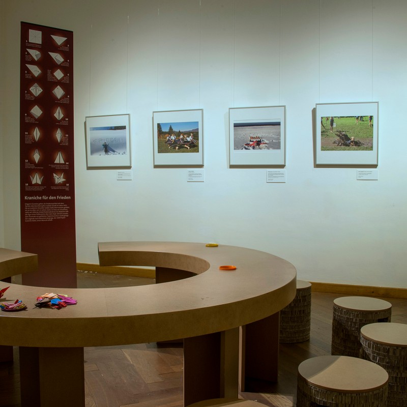 Half-round wooden table with numerous colorful origami figures, a wall displays five framed photographs and a panel
                                 with paper crane folding instructions. (AI-generated alt-text, generated with GPT-4.1-mini)