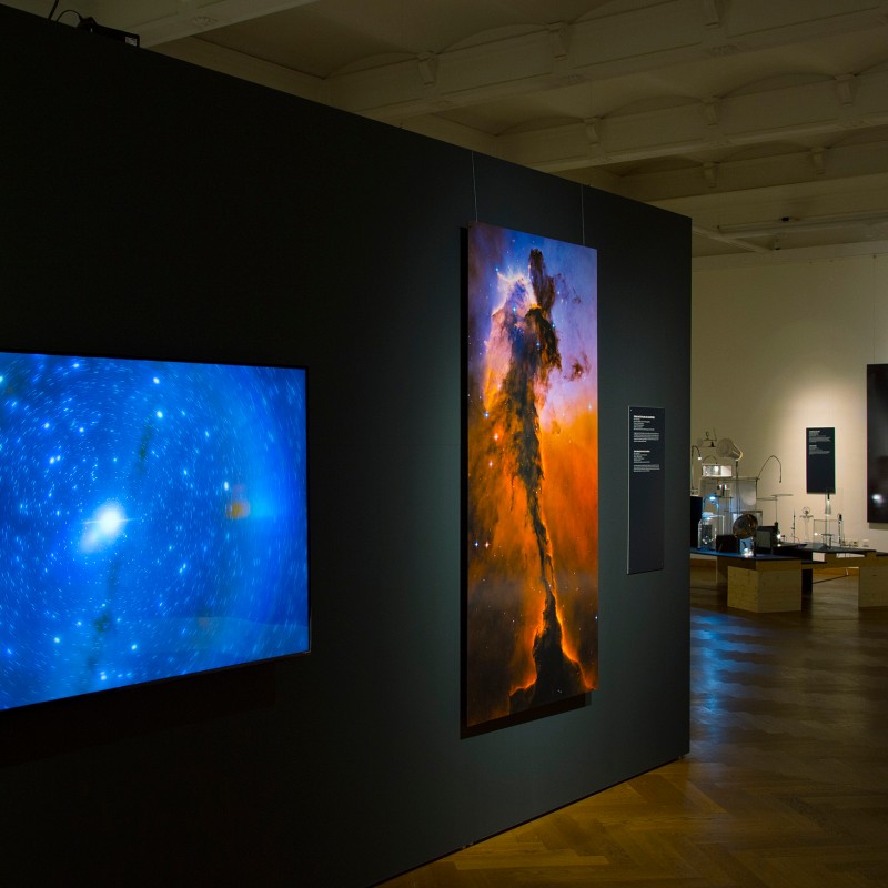 Dimly lit exhibition room with a glowing screen, a vertical image of a space cloud, and additional astronomical
                                 exhibits in the background. (AI-generated alt-text, generated with GPT-4.1-mini)