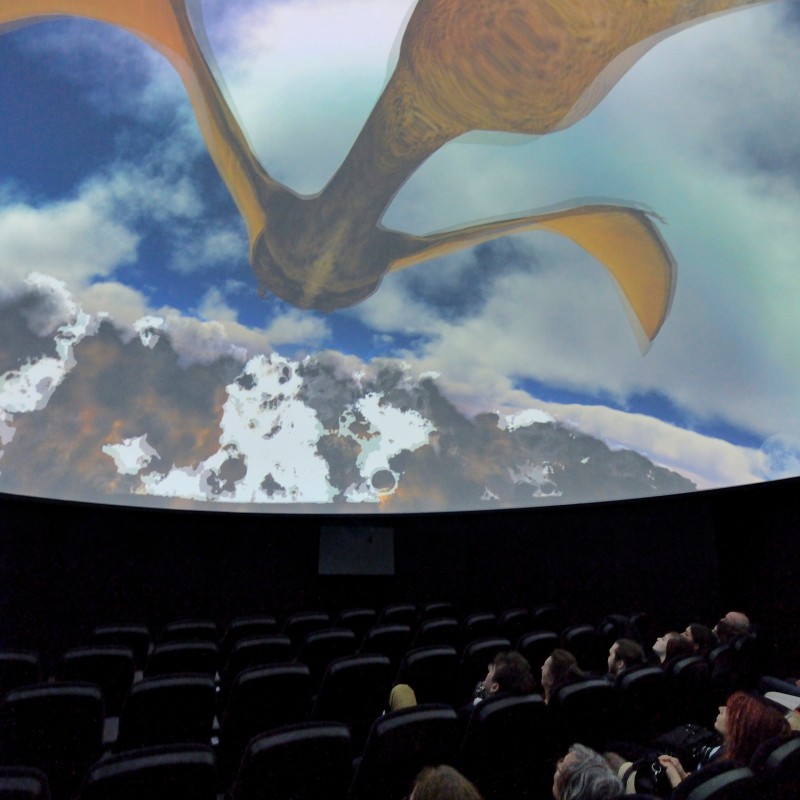 Audience seated in a theater watches a large screen projection of a flying pterosaur above clouds. (AI-generated
                                 alt-text, generated with GPT-4.1-mini)