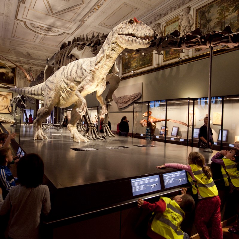 Large dinosaur skeleton and model exhibit in a museum with children in yellow safety vests observing closely (AI-generated
                                 alt-text, generated with GPT-4.1-mini)