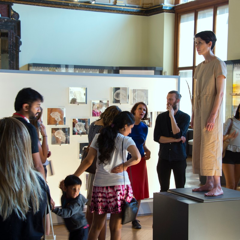 Woman in beige dress stands motionless on a pedestal, surrounded by several adults and children in an exhibition
                                 room. (AI-generated alt-text, generated with GPT-4.1-mini)