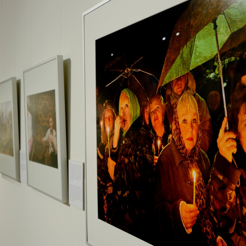 Exhibition view with several framed photos on a white wall, foreground shows a photograph of people holding candles
                                 and umbrellas. (AI-generated alt-text, generated with GPT-4.1-mini)