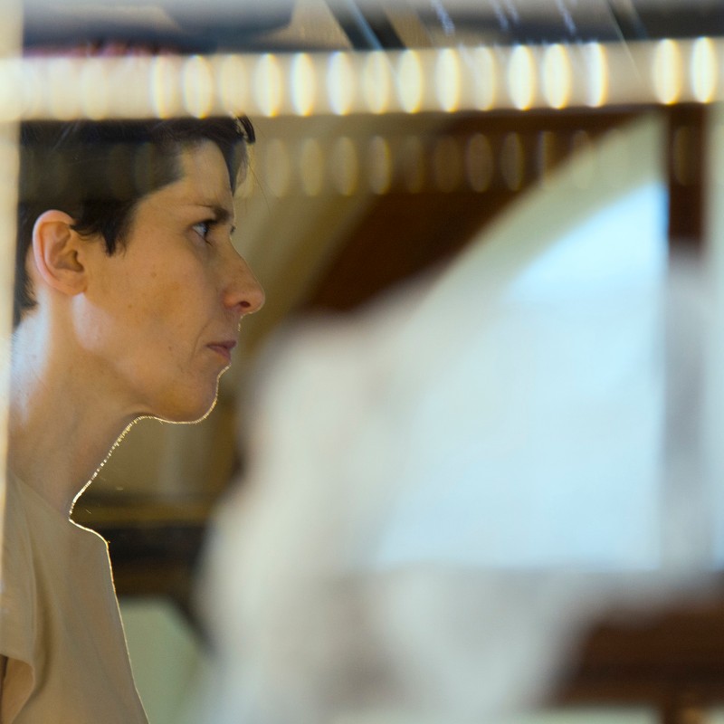 Woman with short dark hair in profile, focused expression, soft lighting, blurred glass reflections in foreground.
                                 (AI-generated alt-text, generated with GPT-4.1-mini)