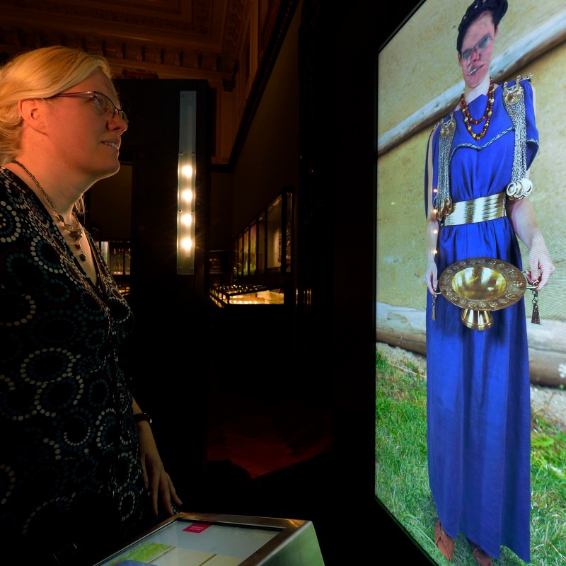 Frau
                                 mit langem, blondem Haar und Brille blickt auf einen großen, stehenden Bildschirm mit Darstellung einer Frau in blauer Robe
                                 und antikem Schmuck. (KI-generierter Alt-Text, erstellt mit GPT-4.1-mini)