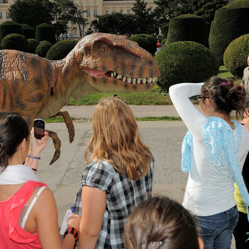 Group of young women on a forecourt looking at and photographing a life-size dinosaur model. Trimmed bushes and
                                 an older building are visible in the background. (AI-generated alt-text, generated with GPT-4.1-mini)
