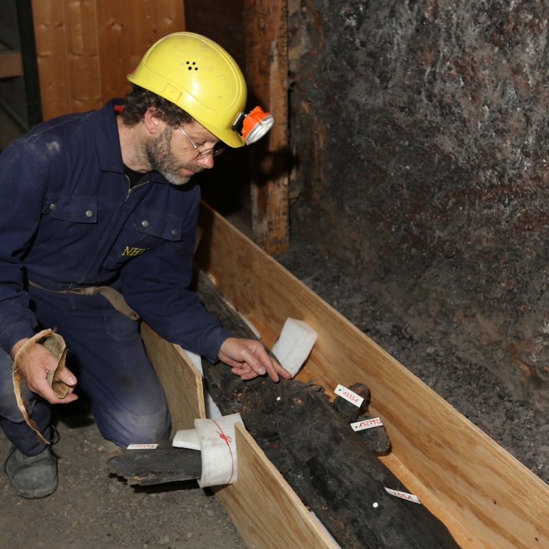 Mann mit Schutzhelm und Stirnlampe in Arbeitskleidung kniet neben Holzfund in einer Kiste; mehrere kleine Schilder
                                 befestigt. (KI-generierter Alt-Text, erstellt mit GPT-4.1-mini)