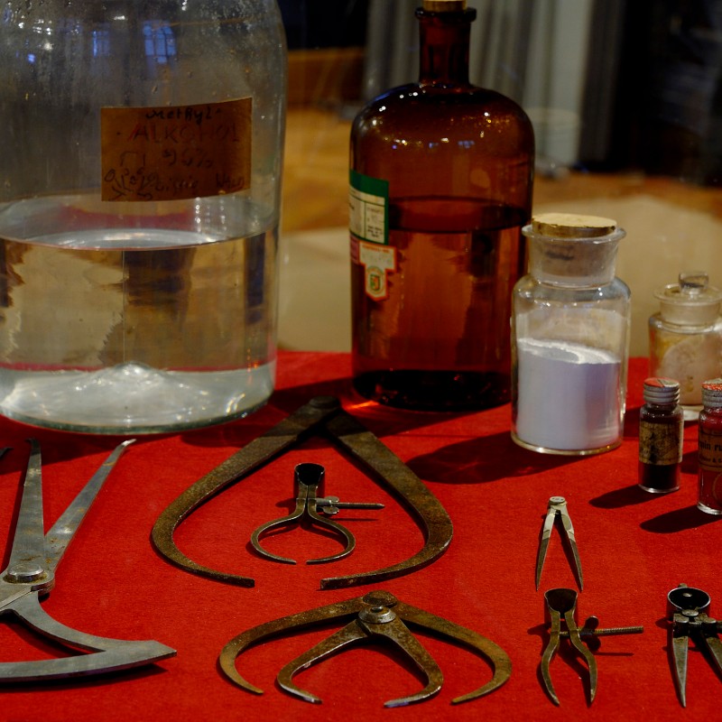 Old metal measuring instruments and various glass containers with liquids and powders on red cloth. (AI-generated
                                 alt-text, generated with GPT-4.1-mini)