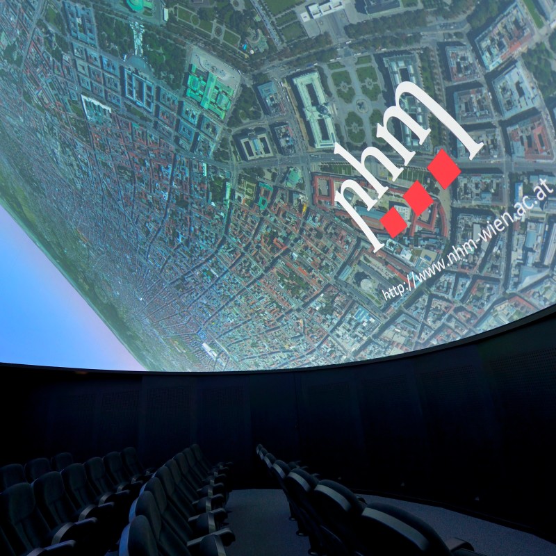 Rows of empty cinema seats facing a large curved screen displaying a city view and nhm logo. (AI-generated alt-text,
                                 generated with GPT-4.1-mini)