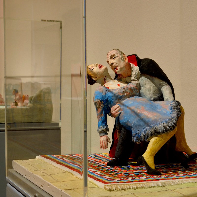 Plastik zweier Figuren: Eine vampirähnliche Gestalt hält eine Frau im blauen Kleid in den Armen, beide auf gemustertem
                                 Teppich. Im Hintergrund weitere Figuren. (KI-generierter Alt-Text, erstellt mit GPT-4.1-mini)