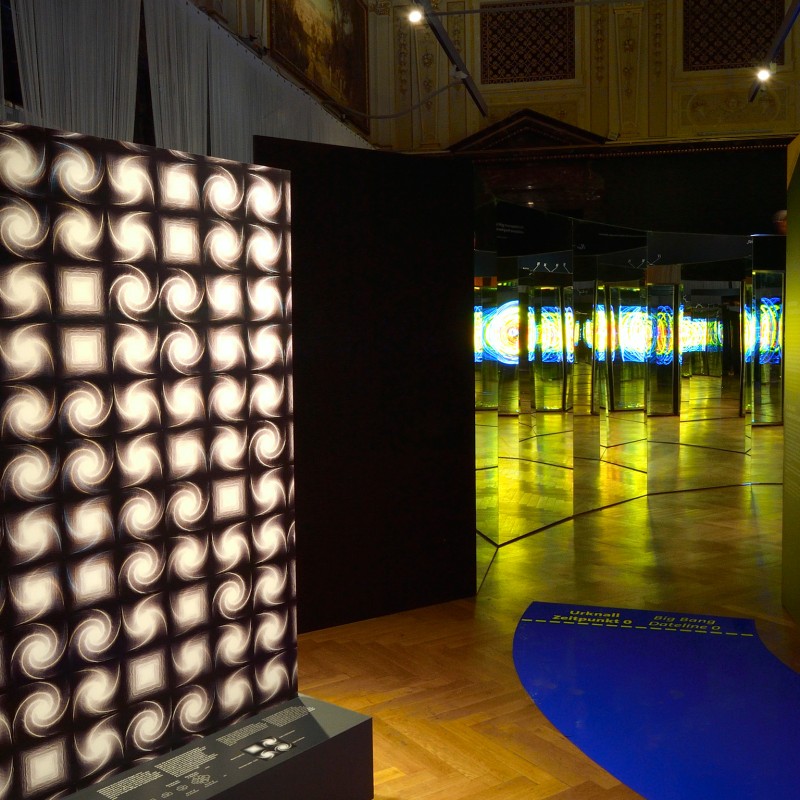 Leuchtwand mit quadratischen Mustern aus Licht und Schatten, im Hintergrund beleuchtete, hochformatige Glasstelen
                                 mit Farbmuster. (KI-generierter Alt-Text, erstellt mit GPT-4.1-mini)