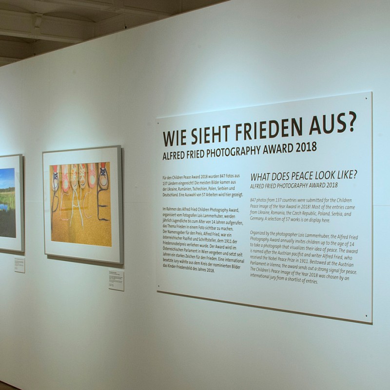 Museum exhibition with photographs on a white wall, central large panel with the heading 'WIE SIEHT FRIEDEN AUS?'
                                 and text below. To the right, a framed photo with a person in the foreground. (AI-generated alt-text, generated with GPT-4.1-mini)