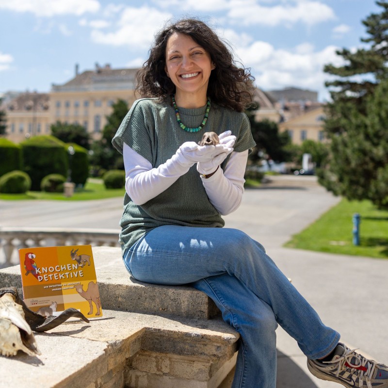 Konstantina Saliari
                                 is sitting in front of the entrance to the Natural History Museum in Vienna, holding the skeleton of a small animal skull
                                 in her hands. Next to her is the book 