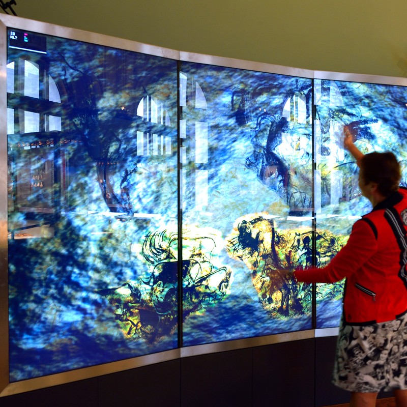 Frau im roten Blazer mit ausgestrecktem Arm vor einer großen, beleuchteten Ausstellungswand mit multicolor Abbildungen.
                                 (KI-generierter Alt-Text, erstellt mit GPT-4.1-mini)