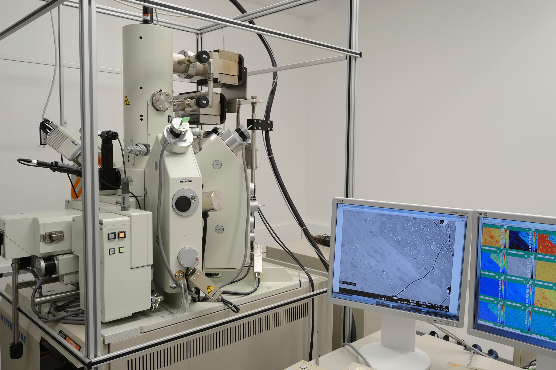 : Large research laboratory instrument with cylindrical and rectangular components, surrounded by a metal frame. Next to it are two monitors showing scientific images and colored grid maps. (AI-generated alt-text, generated with GPT-4.1-mini)