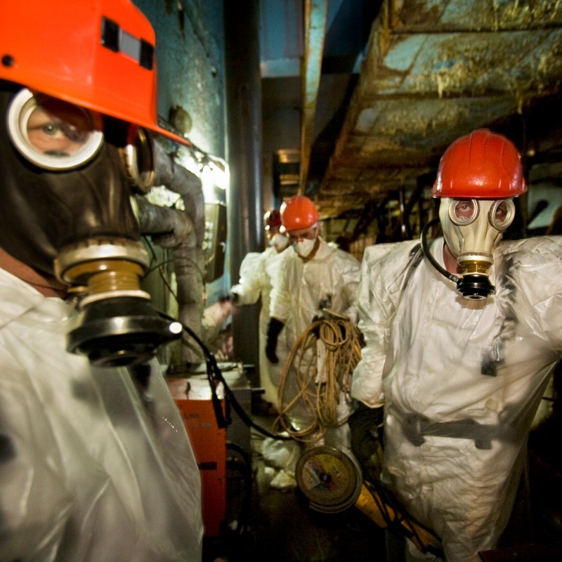 Drei Personen in weißen Schutzanzügen, roten Helmen und Atemschutzmasken stehen in einem Industriegebäude mit Kabeln
                                 und technischen Geräten. (KI-generierter Alt-Text, erstellt mit GPT-4.1-mini)