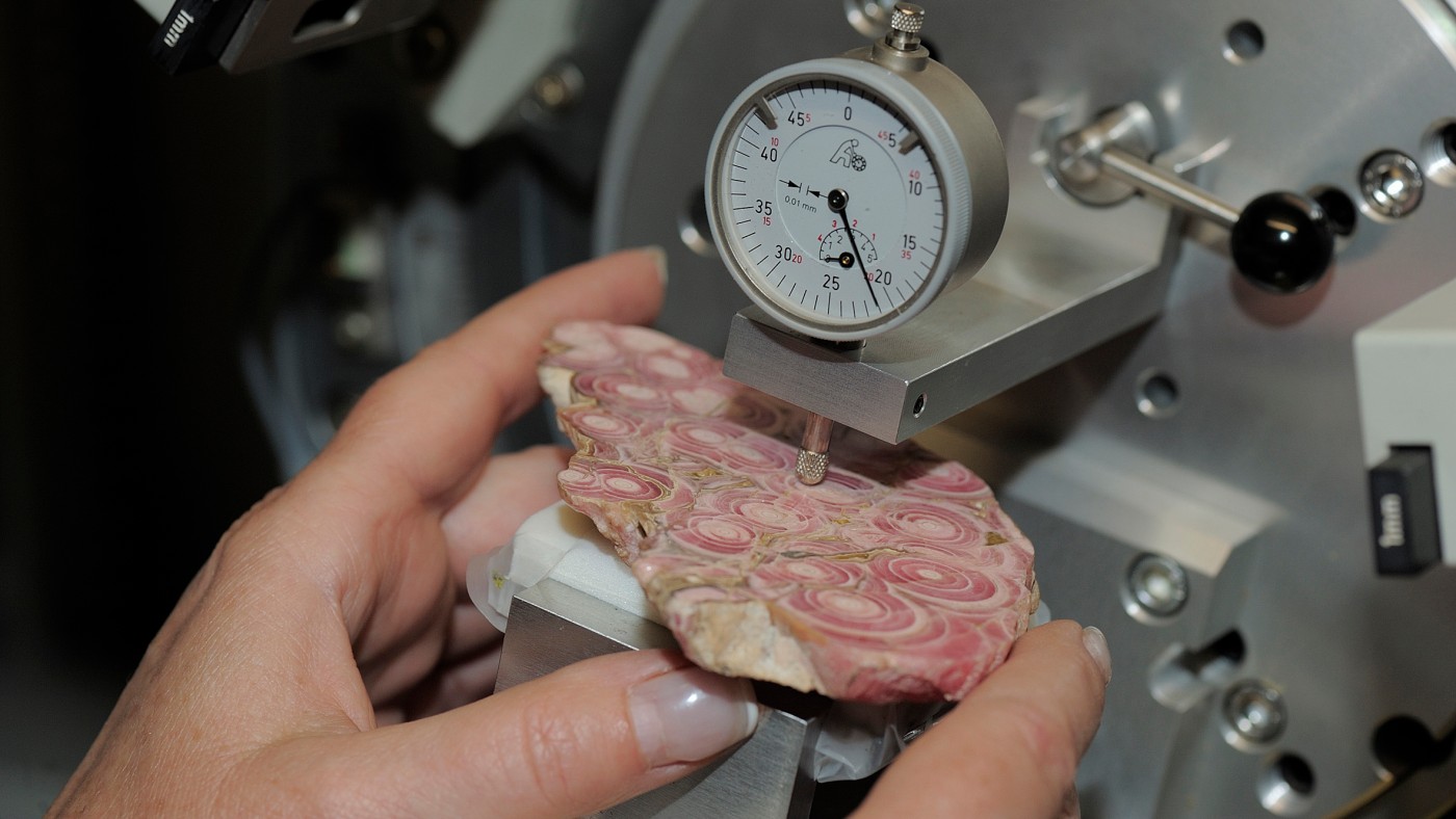 : Nahaufnahme von Händen, die eine polierte Gesteinsplatte mit runden, rosa-grauen Strukturen unter einem Messgerät halten. (KI-generierter Alt-Text, erstellt mit GPT-4.1-mini)