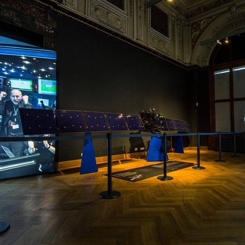 Replik
                                 eines Raumschiffs mit ausgebreiteten Solarpaneelen auf Holzfußboden, im Hintergrund eine große Fotowand mit jubelndem Mann.
                                 (KI-generierter Alt-Text, erstellt mit GPT-4.1-mini)