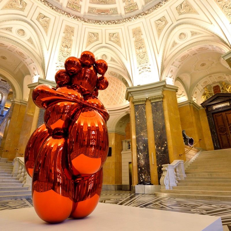 Glossy,
                                 orange-red sculpture with rounded shapes stands in the center of a marble-lined hall with an ornate domed ceiling. (AI-generated
                                 alt-text, generated with GPT-4.1-mini)