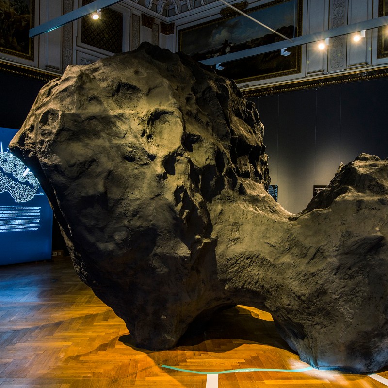 Großes,
                                 dunkelgraues Modell eines unregelmäßig geformten Himmelskörpers mit vielen Vertiefungen steht im Ausstellungsraum mit Parkettboden.
                                 Im Hintergrund Leuchttafel mit weißem Text und Grafiken. (KI-generierter Alt-Text, erstellt mit GPT-4.1-mini)