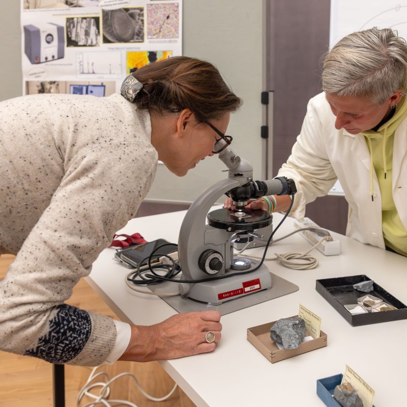 Zwei Personen blicken auf ein graues Mikroskop auf einem Tisch mit Gesteinsproben
                                 und Informationskarten.