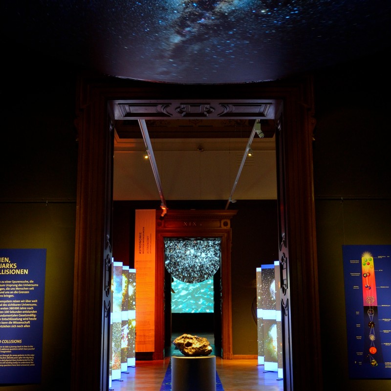 Blick
                                 durch eine offenstehende Tür in einen abgedunkelten Ausstellungsraum mit beleuchteten Vitrinen, zentral liegt ein großer Stein.
                                 An der Decke Projektion einer Galaxie. (KI-generierter Alt-Text, erstellt mit GPT-4.1-mini)