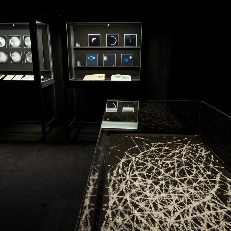 Dark exhibition room with two illuminated display cases and a table with a white, tangled pattern under glass. (AI-generated
                                 alt-text, generated with GPT-4.1-mini)