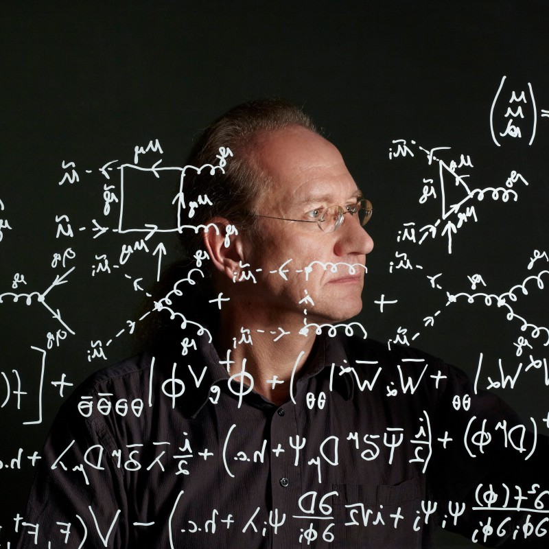 Eine Person mit Brille blickt konzentriert durch eine große Scheibe mit zahlreichen handgeschriebenen physikalischen
                                 Formeln und Diagrammen. (KI-generierter Alt-Text, erstellt mit GPT-4.1-mini)
