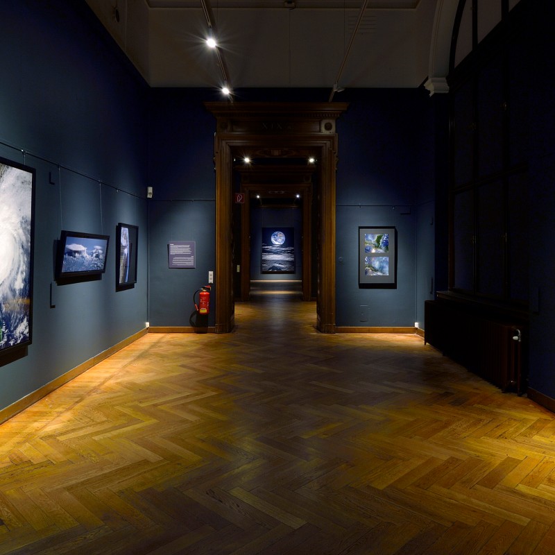 Dark blue exhibition room with large photographs of Earth and space on the walls, wooden floor and view through
                                 an open doorway into another room. (AI-generated alt-text, generated with GPT-4.1-mini)