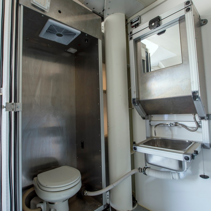 Small cabin with a toilet, stainless steel sink, and mirror; white and gray walls with visible pipes. (AI-generated
                                 alt-text, generated with GPT-4.1-mini)