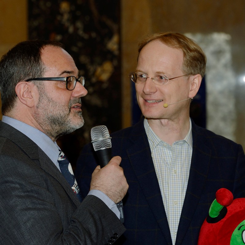 Two men in suits stand side by side. The man on the left speaks into a microphone. Both wear glasses; the man on
                                 the right has a headset. Foreground: Plush figure with red legs and green antennae. (AI-generated alt-text, generated with
                                 GPT-4.1-mini)