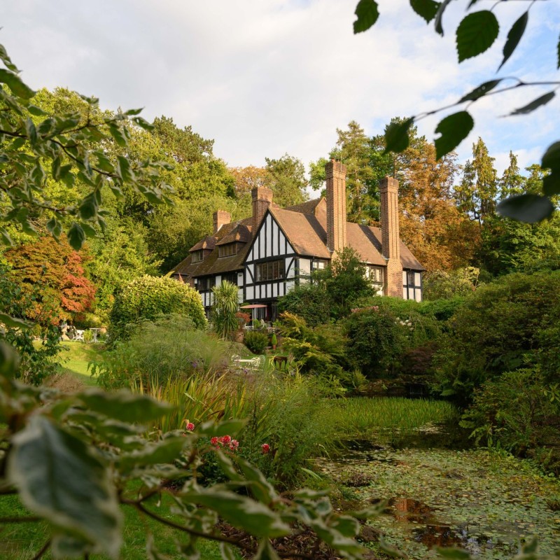 Englisches Landhaus mit Fachwerkfassade und hohen Schornsteinen, umgeben von dichtem, grünem Garten mit Teich. (KI-generierter
                                 Alt-Text, erstellt mit GPT-4.1-mini)