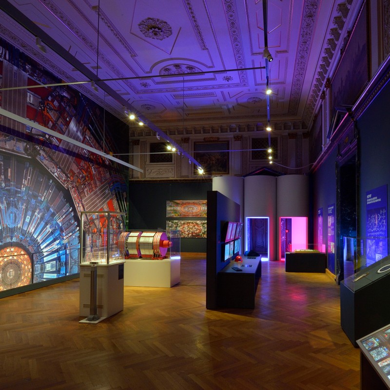 Großer
                                 Ausstellungsraum mit verzierten Stuckdecken, birnenförmigem Holzparkett, Wandprojektionen, beleuchteten Vitrinen und Informationstafeln.
                                 (KI-generierter Alt-Text, erstellt mit GPT-4.1-mini)