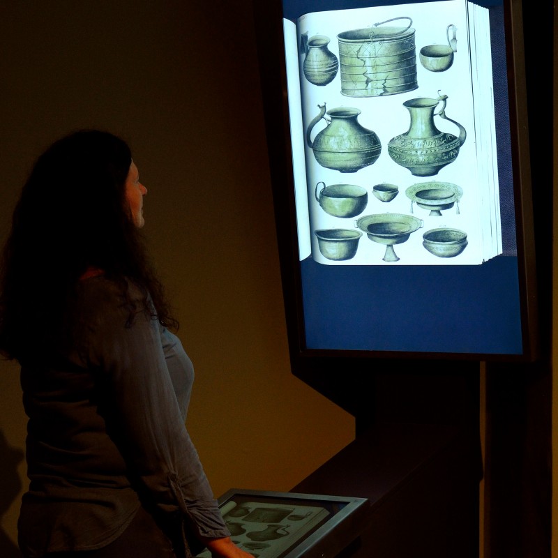 Frau
                                 mit dunklem, lockigem Haar betrachtet ein hochformatiges Display mit einer schwarz-weißen Zeichnung von Gefäßen und Schalen.
                                 (KI-generierter Alt-Text, erstellt mit GPT-4.1-mini)