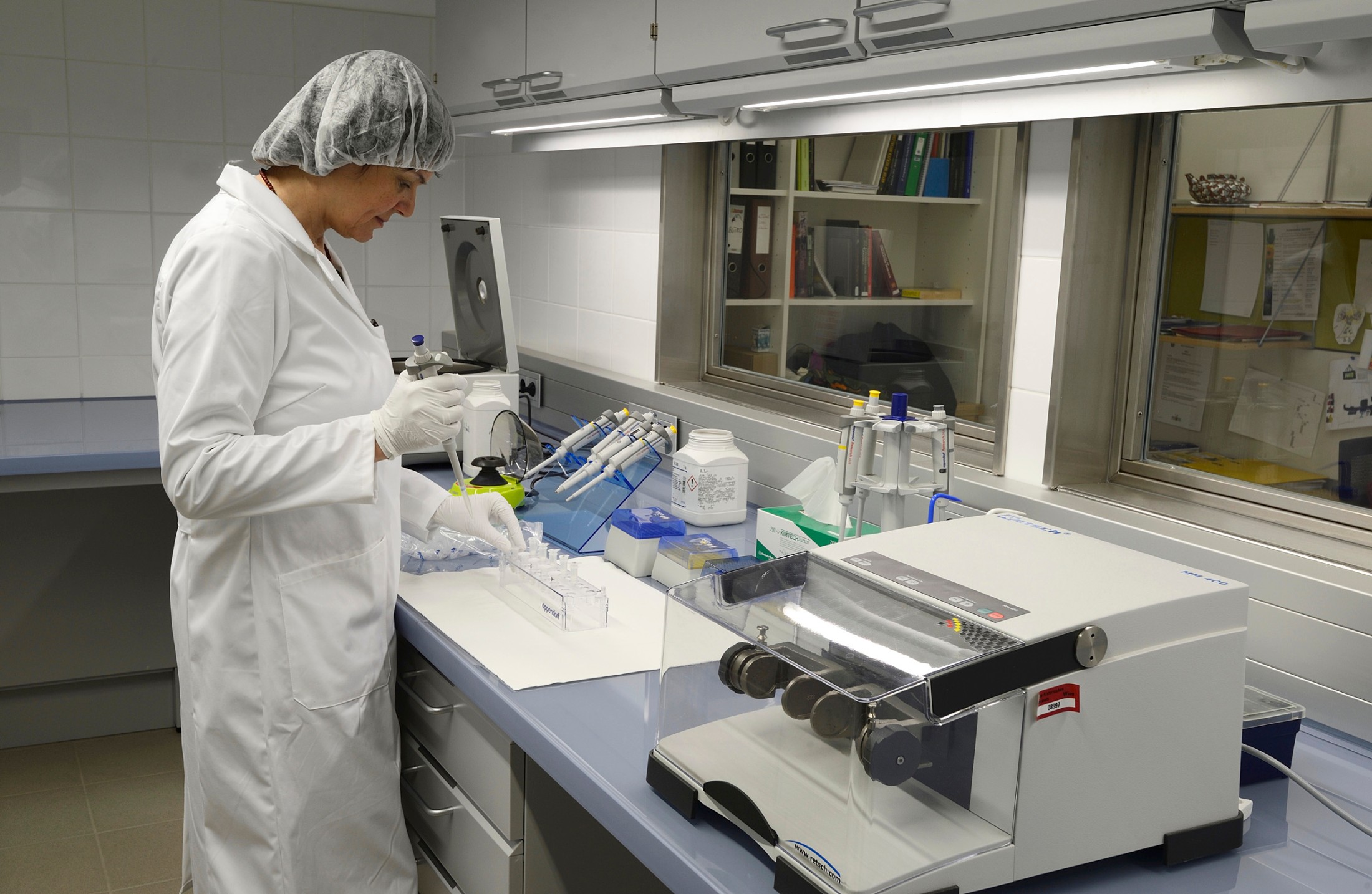 Cleanroom: Person in a white lab coat and hairnet works at a laboratory bench with gloves. Several devices, reagent bottles, and pipettes are present. (AI-generated alt-text, generated with GPT-4.1-mini)