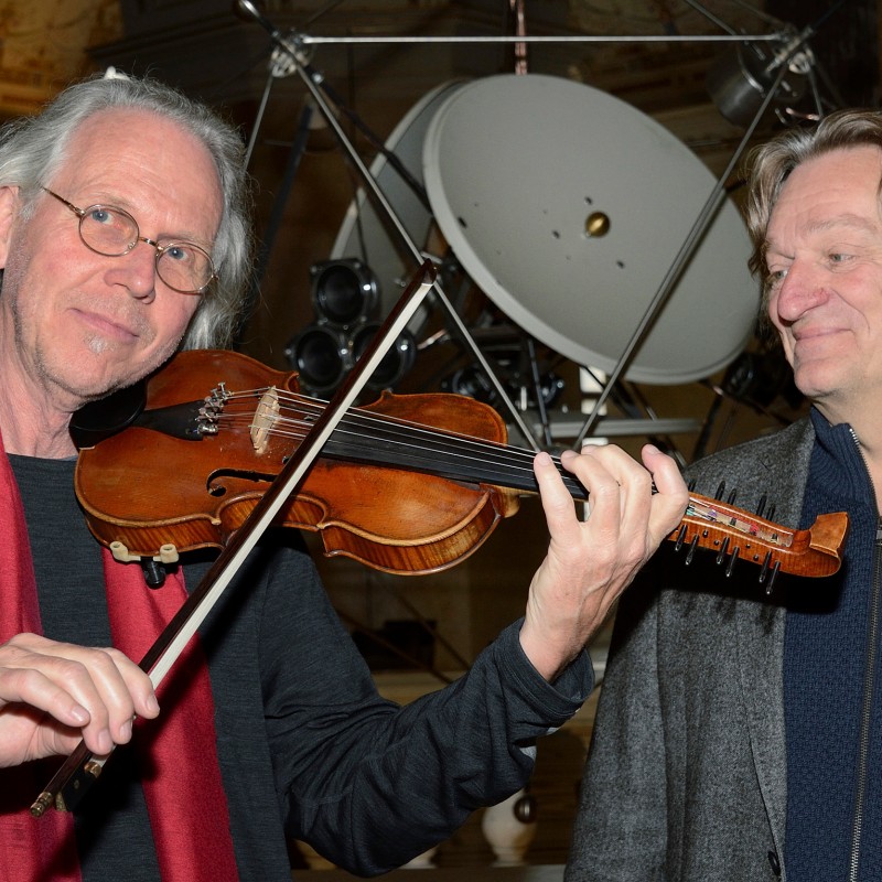 A gray-haired man plays the violin and wears a red scarf, while another blond-haired man stands next to him. In
                                 the background is a large technical device with a parabolic reflector. (AI-generated alt-text, generated with GPT-4.1-mini)